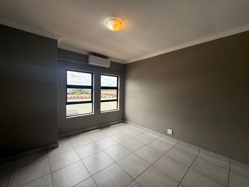 A clean, empty bedroom featuring neutral tiled flooring, an installed air conditioning unit, and large windows providing natural light.