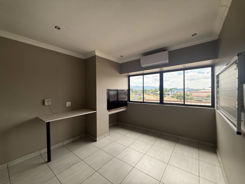 A bright, empty living area featuring tiled floors, a built-in braai, air conditioning, and a wall-mounted desk with a pleasant exterior view.
