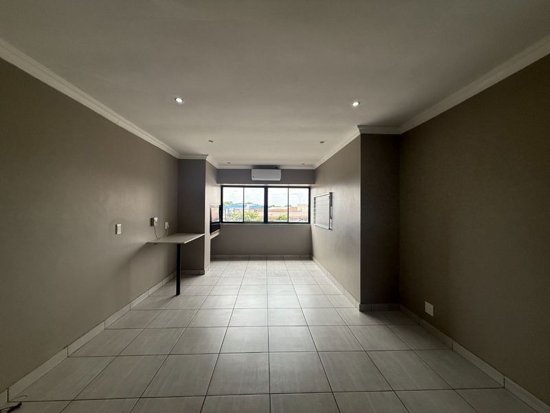 A spacious, empty living area featuring neutral tiled floors, recessed lighting, a wall-mounted air conditioning unit, and a convenient built-in desk space.