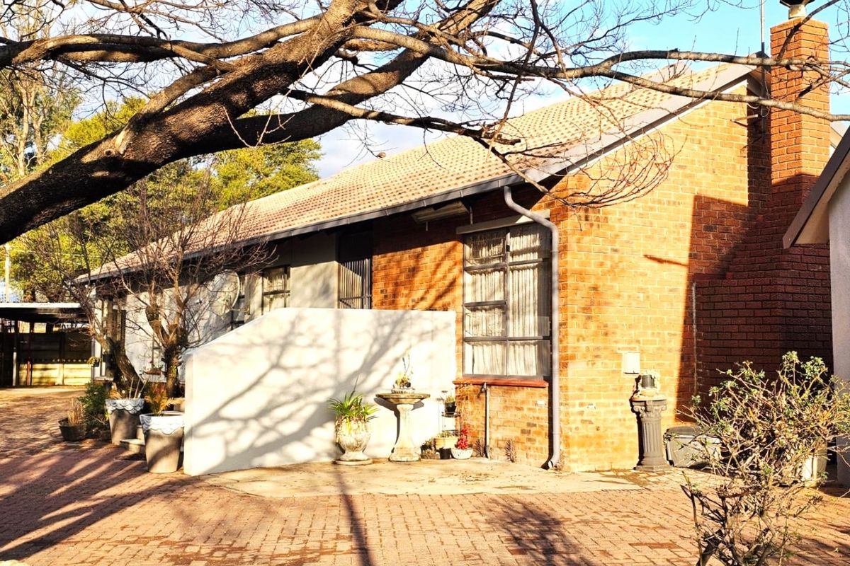 Rear view of a brick house featuring a prominent chimney, tiled roof, and paved outdoor area surrounded by mature trees.