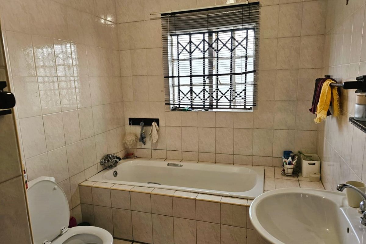 A functional bathroom featuring a built-in bathtub, pedestal basin, and toilet, finished with full-height wall tiling and a security-grilled window.