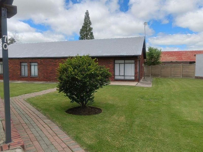 A well-maintained rear exterior view featuring a brick facade, tiled roof, paved walkway, and a spacious lawn area with timber fencing.