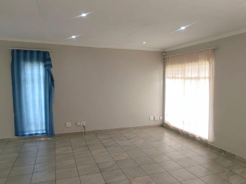 A spacious, empty living area featuring neutral tiled flooring, recessed ceiling downlights, and large windows with installed curtain rails.