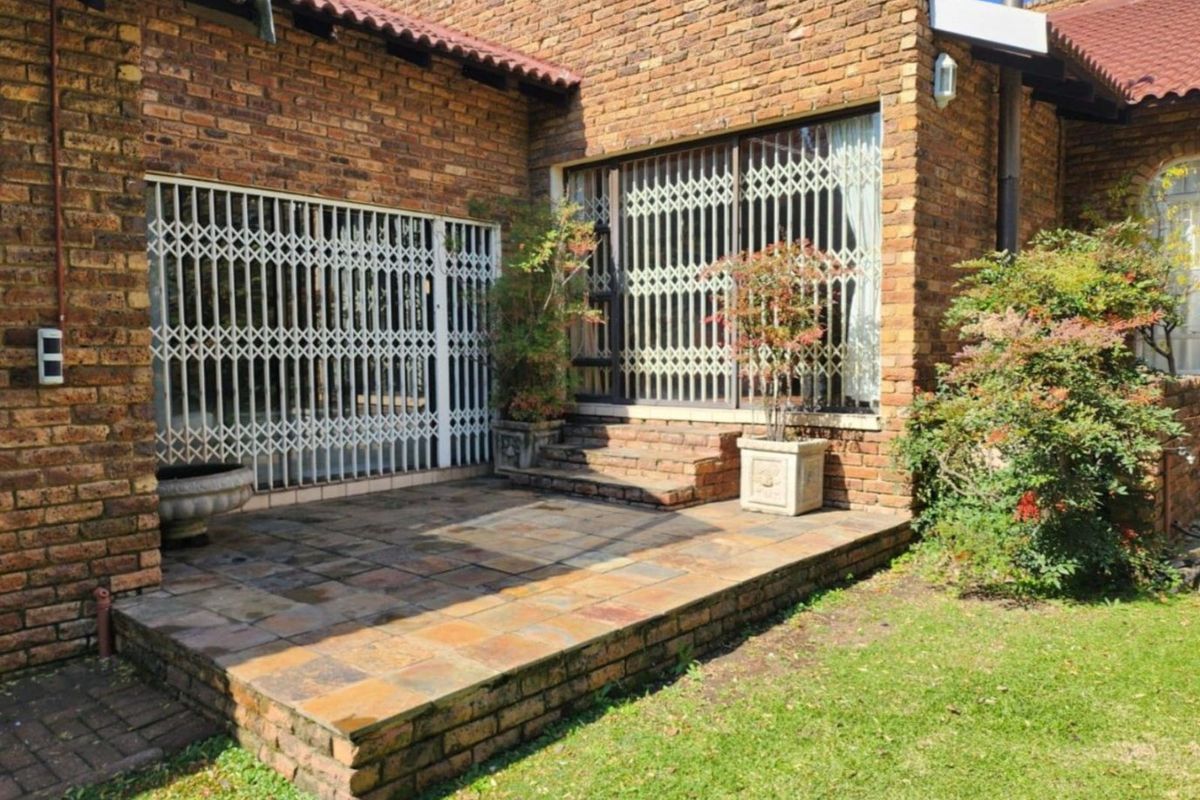 A rear patio area featuring exposed brick walls, tiled flooring, and security gates leading into the home, set against a small garden.