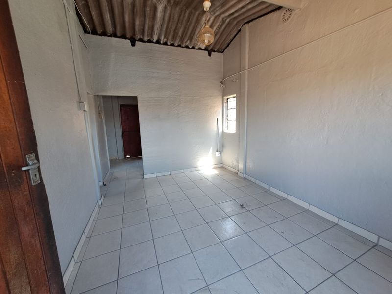 An empty living area featuring tiled flooring, exposed corrugated ceiling, and a simple window, offering a blank canvas for renovation.