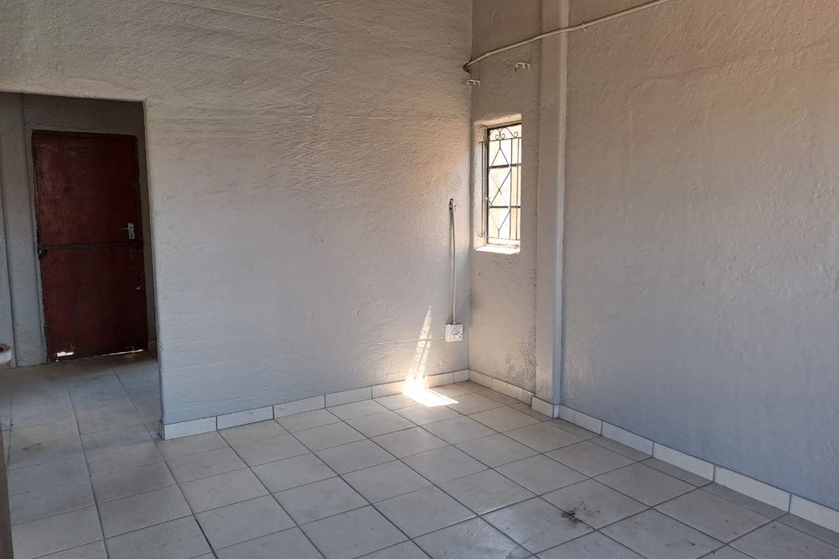 An empty, basic living area featuring ceramic floor tiles, exposed electrical conduit, and a window with security bars.