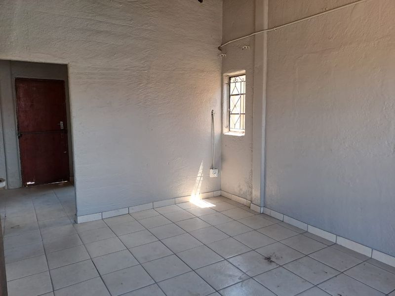 An empty, basic living area featuring ceramic floor tiles, exposed electrical conduit, and a window with security bars.