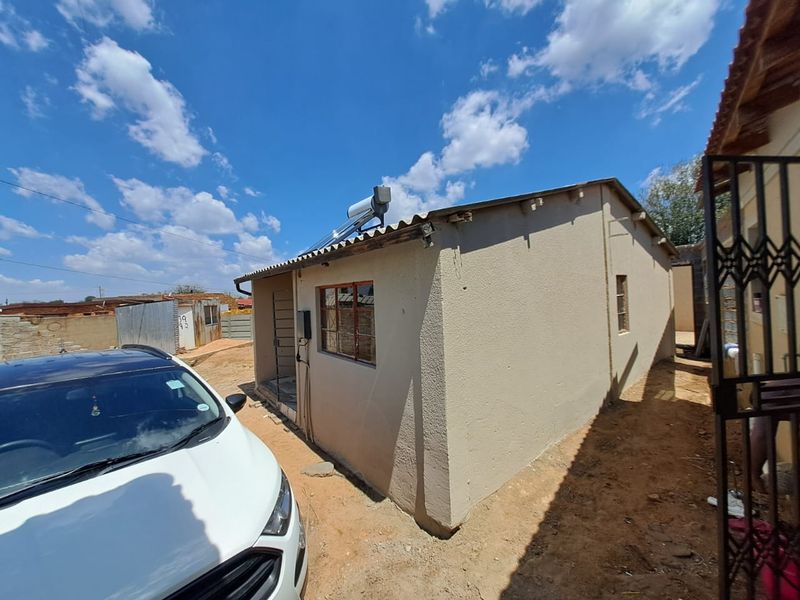 Rear exterior view of a single-story residential dwelling featuring a solar water heating system and corrugated roof sheeting.