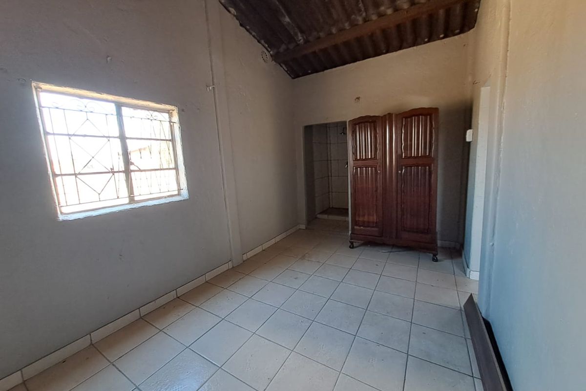 A basic bedroom featuring tiled flooring, exposed rafter ceiling, and a window with security bars, requiring cosmetic updates to improve its overall condition.