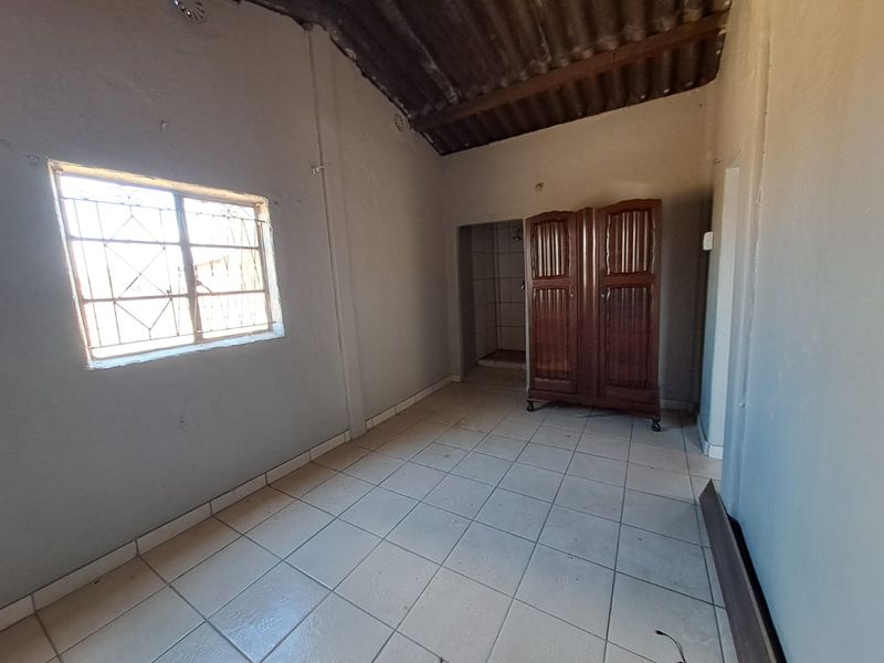 A basic bedroom featuring tiled flooring, exposed rafter ceiling, and a window with security bars, requiring cosmetic updates to improve its overall condition.