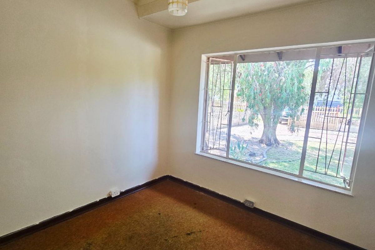 An empty bedroom featuring a large window with security bars, a pendant light fixture, and worn carpet flooring.