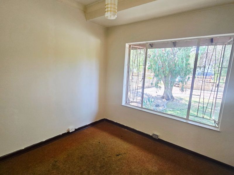 An empty bedroom featuring a large window with security bars, a pendant light fixture, and worn carpet flooring.