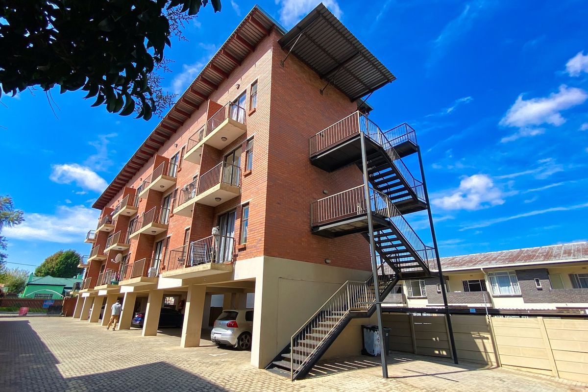 Exterior view of a multi-story apartment building featuring face brick construction, external steel staircases, private balconies, and a paved ground-level parking area.