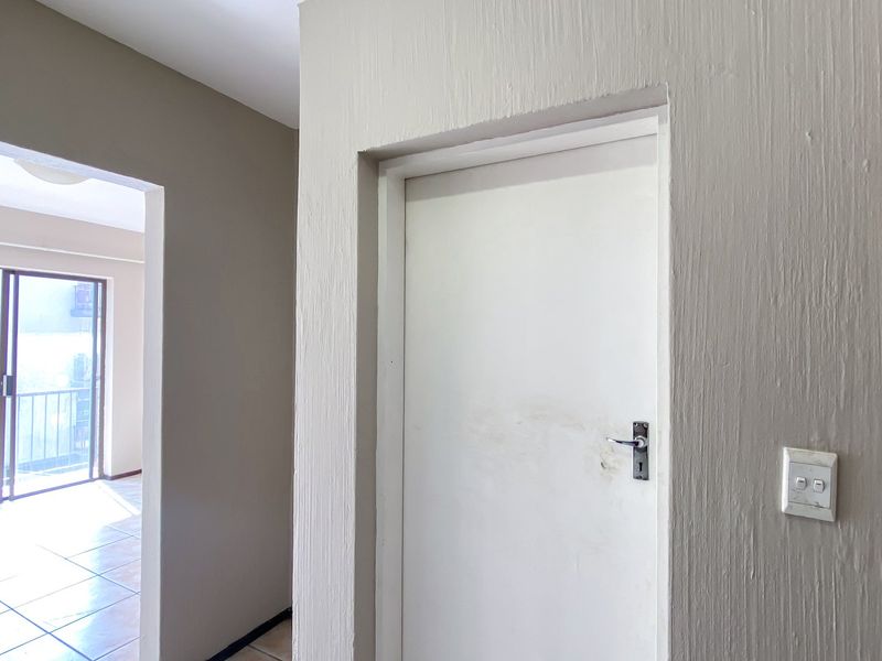 A neutral-toned hallway featuring a standard internal door, light switch, and tiled flooring, leading through to an adjacent living area with balcony access.