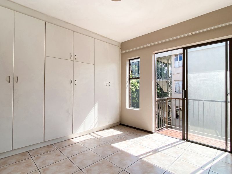 A bright, empty bedroom featuring built-in cupboards, tiled flooring, and a sliding glass door providing access to a private balcony.
