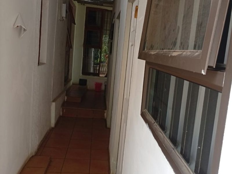 A narrow, tiled hallway featuring exposed roof structure and aluminium windows with security bars, providing access to adjacent rooms.