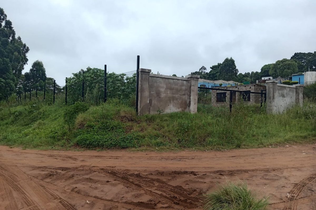 A view of a vacant residential plot featuring perimeter fencing and an unfinished concrete boundary wall, accessible via an unpaved road.