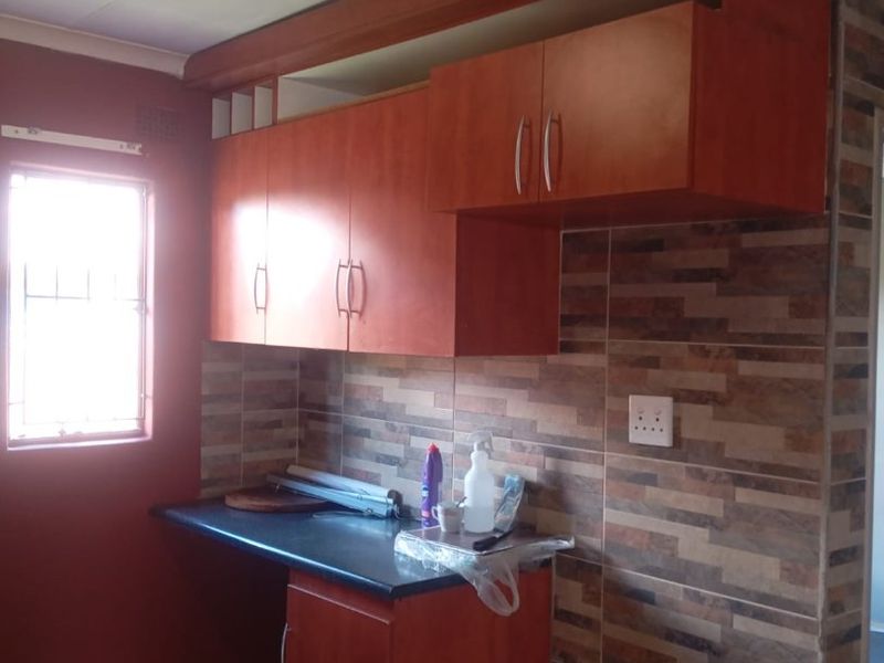 A compact kitchen area featuring built-in wooden cabinetry, a dark laminate countertop, and decorative tiled splashback walls.