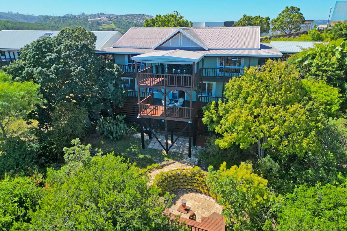 Aerial view of the rear elevation featuring a multi-level timber deck, corrugated metal roofing, and a landscaped garden with a paved patio area.