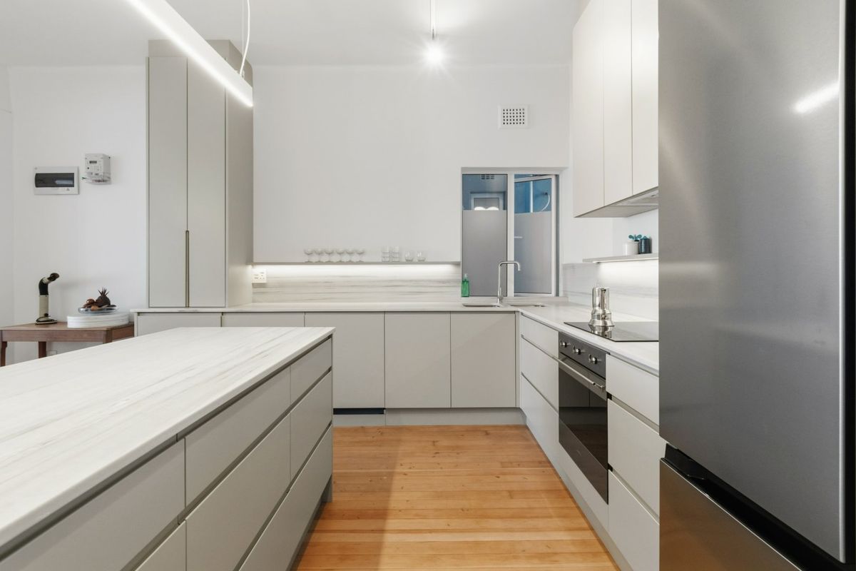 Modern kitchen featuring sleek handleless cabinetry, stone countertops, a central island, built-in appliances, and warm timber flooring.