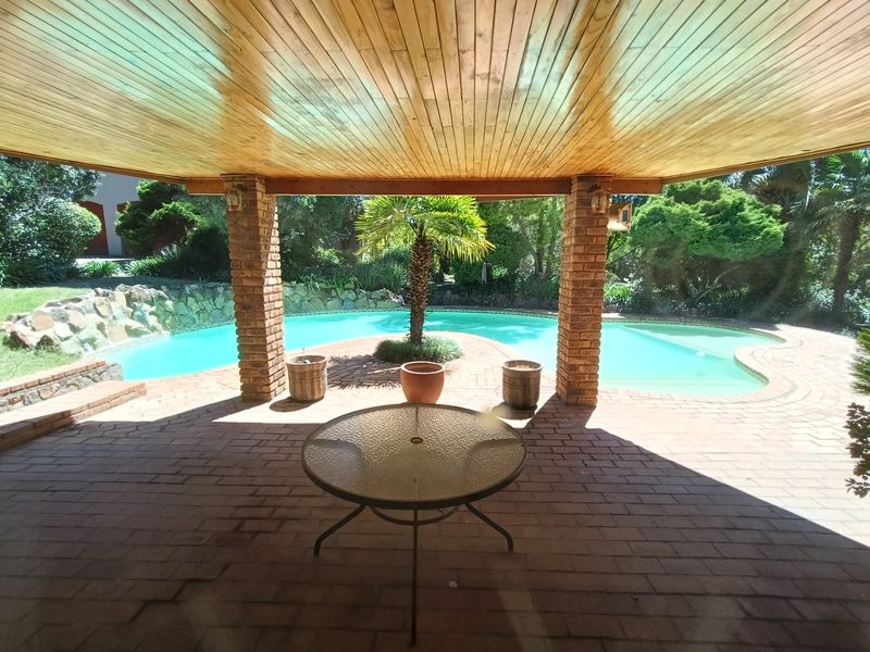 A covered outdoor patio featuring a timber-clad ceiling and brick pillars, overlooking a swimming pool and landscaped garden area.