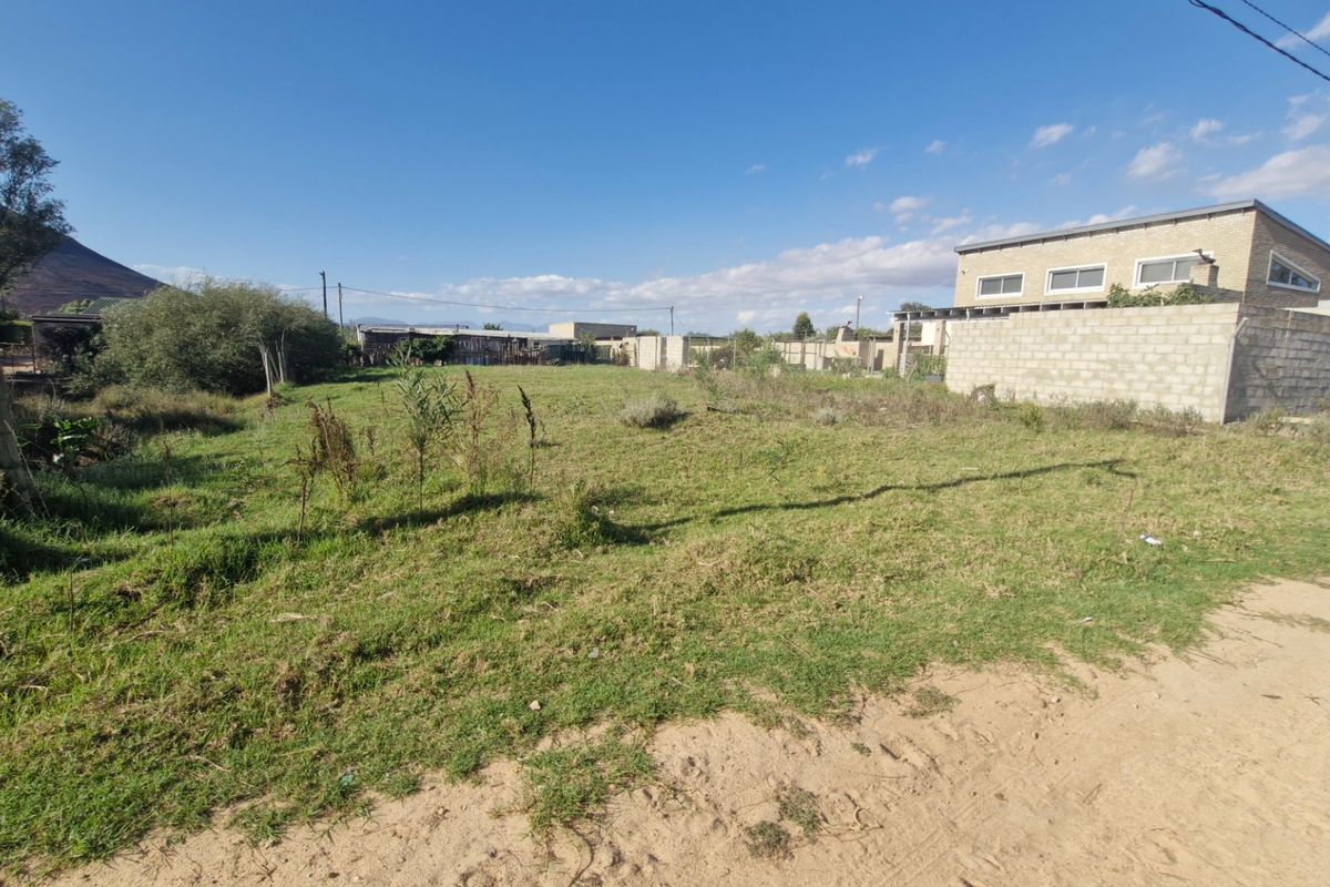 A vacant residential plot featuring grass cover, boundary walling, and direct access from an unpaved road.
