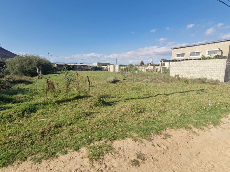 A vacant residential plot featuring grass cover, boundary walling, and direct access from an unpaved road.
