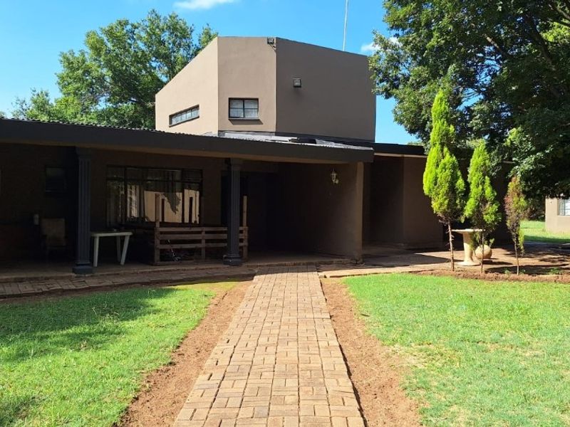 A rear view of the property featuring a paved walkway leading to a covered patio area, surrounded by a lawn and mature trees.