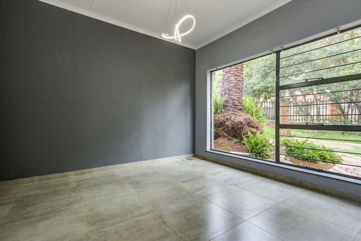 A bright, empty living area featuring large windows with security bars, modern grey tiled flooring, and a neutral dark grey accent wall.