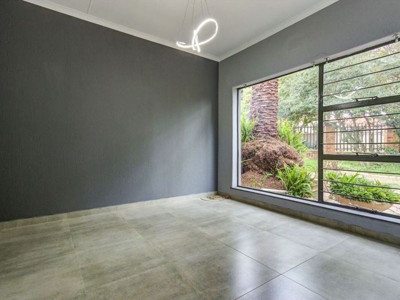 A bright, empty living area featuring large windows with security bars, modern grey tiled flooring, and a neutral dark grey accent wall.
