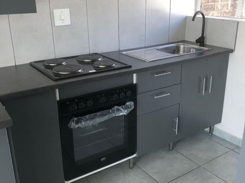 A modern, compact kitchen featuring grey cabinetry, a built-in oven with electric hob, stainless steel sink, and neutral tiled finishes.
