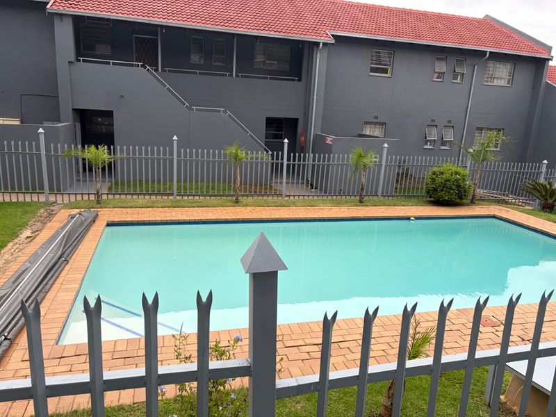 A communal swimming pool area featuring brick coping, a pool cover, and secure metal perimeter fencing set against a multi-story apartment building.