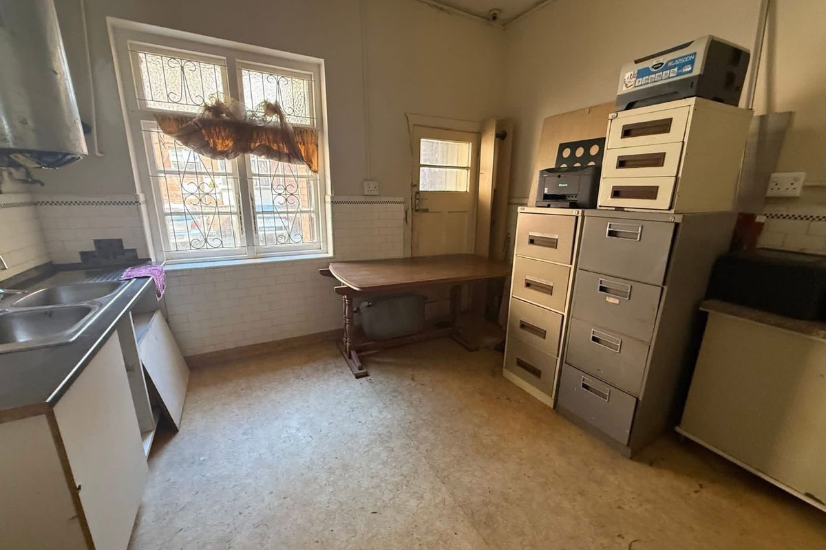 A dated kitchen area featuring a double sink, tiled splashbacks, and a window with security bars, currently used as a utility or storage space.