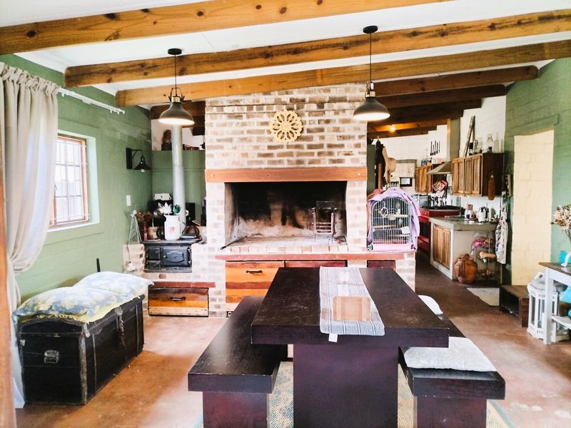 Rustic open-plan living and kitchen area featuring exposed timber beams, a large brick fireplace with integrated wood-burning stove, and concrete flooring.