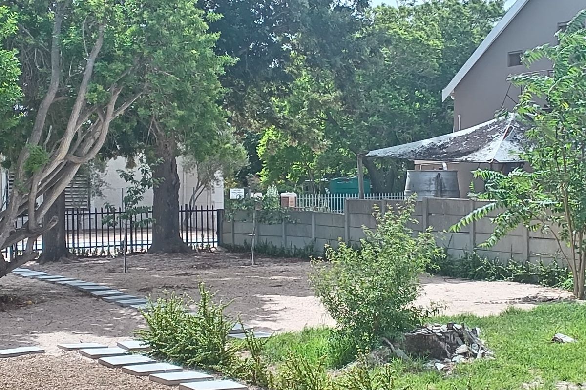 A rear garden area featuring a precast concrete boundary wall, metal fencing, paved stepping stones, and a weathered shade structure with a water tank.