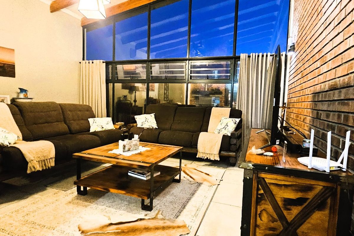 Spacious living room featuring an exposed brick wall, high timber-beamed ceilings, and a large glass window wall providing ample natural light.