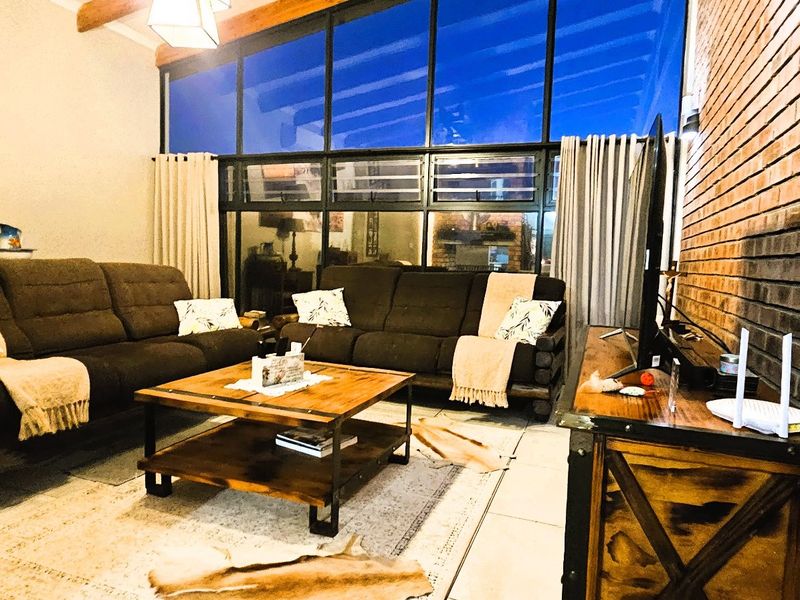 Spacious living room featuring an exposed brick wall, high timber-beamed ceilings, and a large glass window wall providing ample natural light.