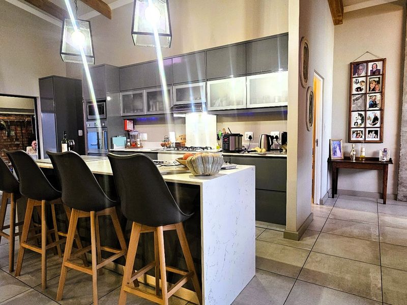 Modern open-plan kitchen featuring a central island with stone countertops, sleek cabinetry, and exposed timber beams, seamlessly flowing into the living area with brick fireplace.
