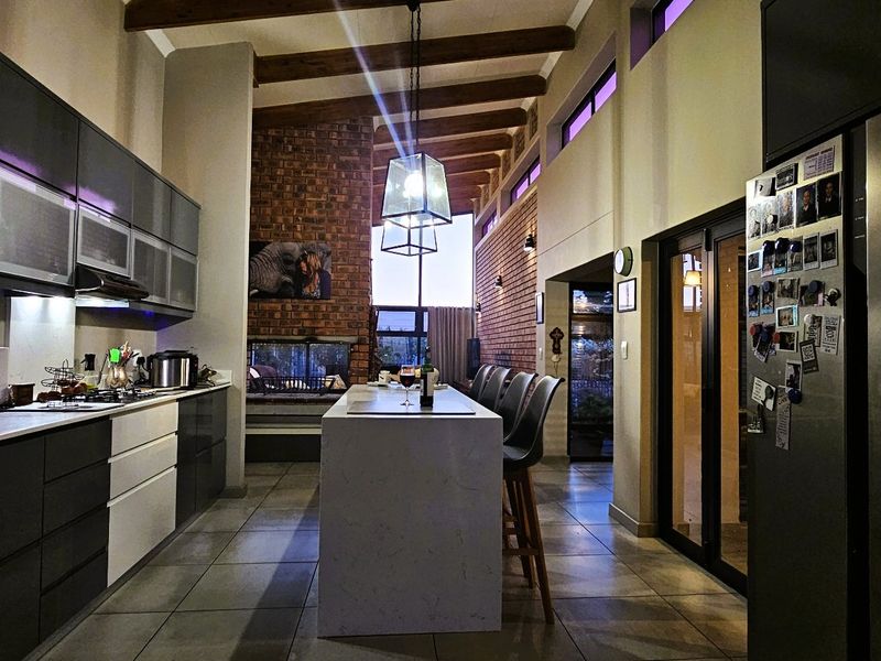 Modern open-plan kitchen featuring a central island, built-in appliances, and a striking brick fireplace with exposed timber beams in a high-ceiling space.