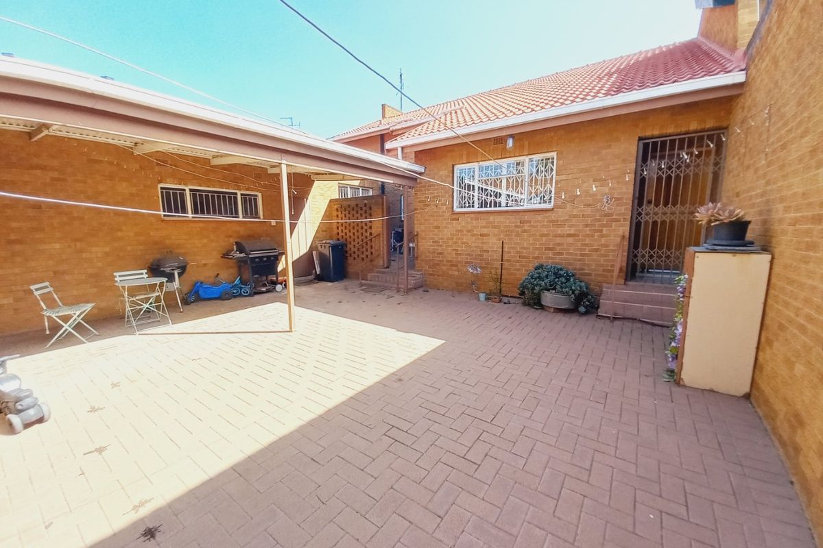 A paved rear courtyard area featuring a covered patio section, brick exterior walls, and secure access points with security gates and window bars.
