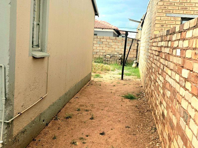 A narrow side or rear exterior passage featuring a plastered house wall, a brick boundary wall, and an unpaved ground area.