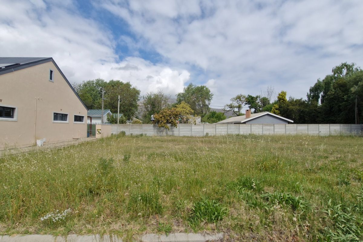 A vacant residential plot featuring a large grassed area, enclosed by a precast concrete boundary wall, adjacent to an existing residential structure.