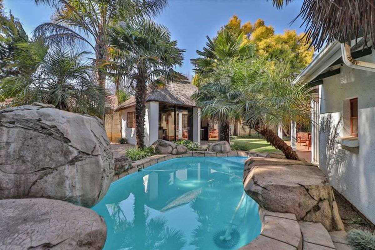 A landscaped backyard featuring a rock-lined swimming pool and a separate thatched-roof outdoor entertainment structure.