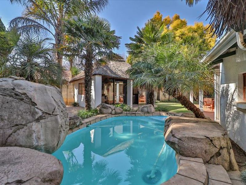A landscaped backyard featuring a rock-lined swimming pool and a separate thatched-roof outdoor entertainment structure.