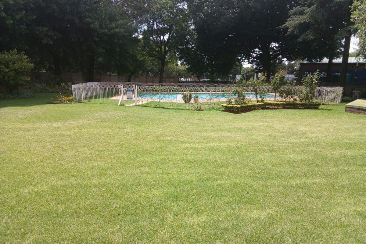 A well-maintained communal garden area featuring a fenced swimming pool surrounded by mature trees and manicured lawn.