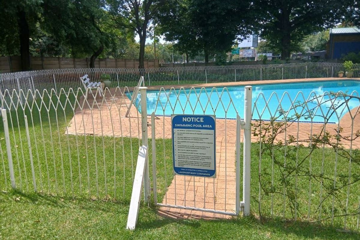 Communal swimming pool area featuring a paved surround, lawn, and secure metal perimeter fencing.