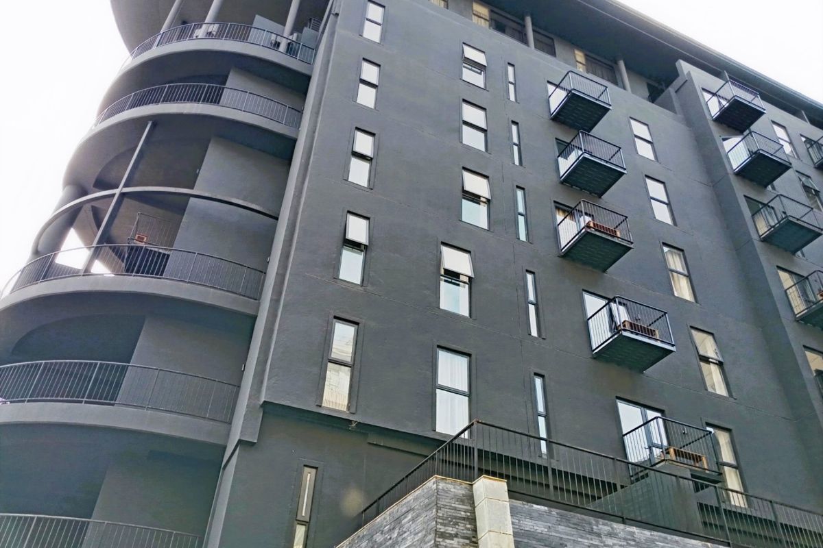 A modern apartment building exterior featuring dark facade finishes, multiple private balconies with metal railings, and stone cladding accents at the base.