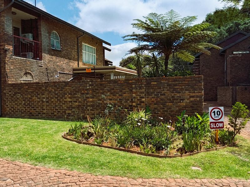 Exterior view of a face brick townhouse complex featuring a paved driveway, landscaped garden, and balcony with metal railings.