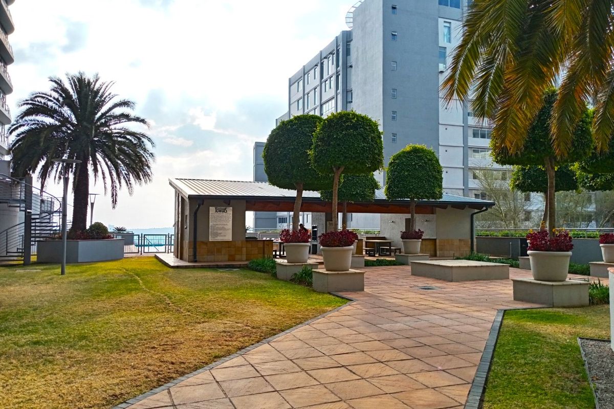 A well-maintained communal outdoor area featuring a paved walkway, landscaped garden, and a covered entertainment structure with stone cladding.
