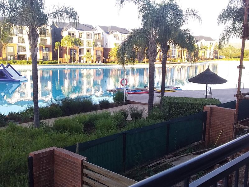 View of a large communal swimming pool and landscaped grounds from an apartment balcony, featuring brick boundary walls and metal railings.